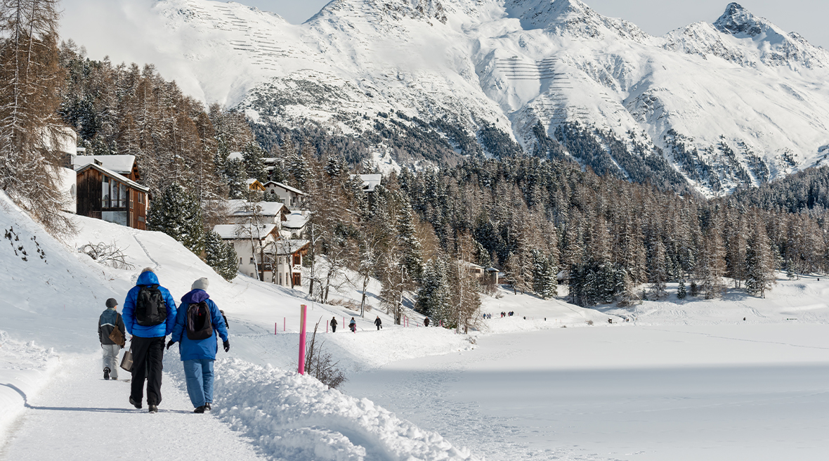 Couple in ski gear walk a snowy path towards mountains and wooden lodges